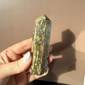 Puede incluir: Una gema pulida y columnar con un patr&oacute;n moteado de verde, marr&oacute;n y blanco. El cristal se sostiene en una mano, mostrando sus tonos naturales y terrosos y su textura &uacute;nica. La parte superior es una superficie plana y angular.
