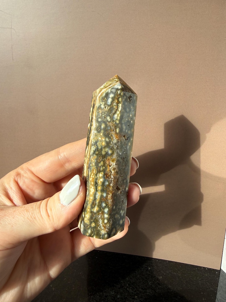 Puede incluir: Una gema pulida y columnar con un patr&oacute;n moteado de verde, marr&oacute;n y blanco. El cristal se sostiene en una mano, mostrando sus tonos naturales y terrosos y su textura &uacute;nica. La parte superior es una superficie plana y angular.