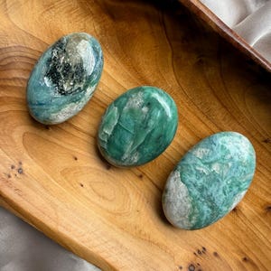 Puede incluir: Tres piedras pulidas de forma ovalada y superficie lisa en varios tonos de verde y blanco. Cada piedra presenta un veteado natural único, texturas cristalinas y acabados brillantes. Las piedras están dispuestas en diagonal, mostrando patrones distintos de verde bosque profundo, tonos menta más claros e inclusiones minerales blancas opacas en sus formas redondeadas.