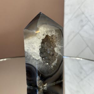 May include: A polished, gray and white agate crystal tower with a hollowed-out center revealing a sparkling geode. The crystal is displayed on a reflective surface, showcasing its natural patterns and textures. The crystal is a decorative item.