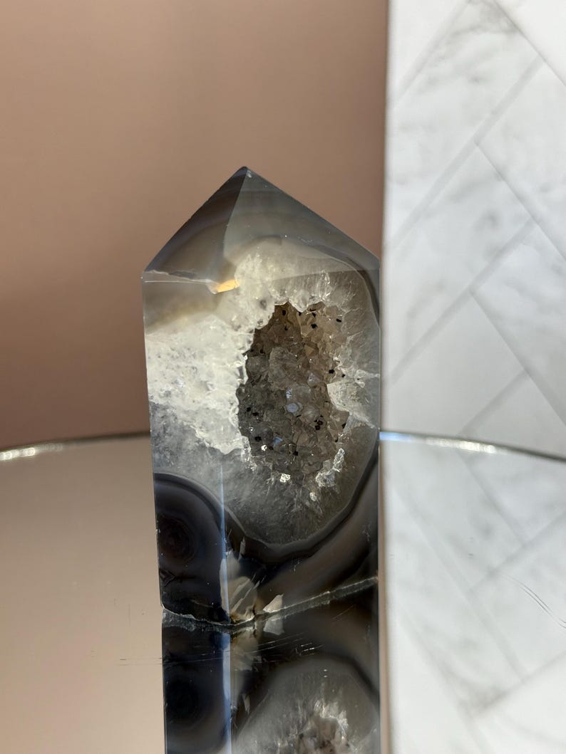 May include: A polished, gray and white agate crystal tower with a hollowed-out center revealing a sparkling geode. The crystal is displayed on a reflective surface, showcasing its natural patterns and textures. The crystal is a decorative item.