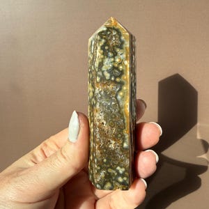 Puede incluir: Un obelisco de jaspe oce&aacute;nico pulido, con una mezcla de colores verdes, marrones y crema con un patr&oacute;n moteado. La piedra preciosa es rectangular con una parte superior puntiaguda. El obelisco se sostiene en una mano.