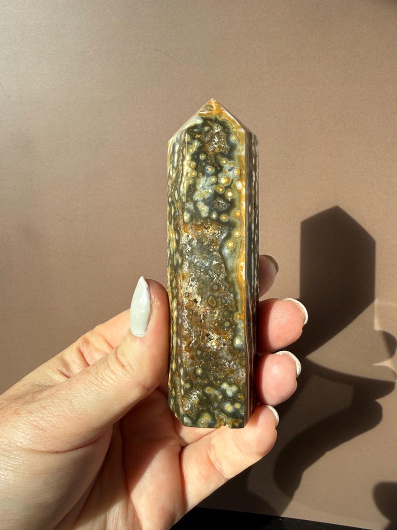 Puede incluir: Un obelisco de jaspe oce&aacute;nico pulido, con una mezcla de colores verdes, marrones y crema con un patr&oacute;n moteado. La piedra preciosa es rectangular con una parte superior puntiaguda. El obelisco se sostiene en una mano.