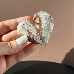May include: A heart-shaped stone with a unique pattern of gray, white, and reddish-brown swirls. The polished surface of the stone reflects light, highlighting the natural variations in color and texture. The stone is held in a hand.