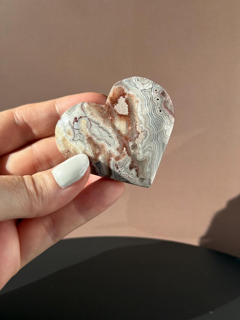 May include: A heart-shaped stone with a unique pattern of gray, white, and reddish-brown swirls. The polished surface of the stone reflects light, highlighting the natural variations in color and texture. The stone is held in a hand.