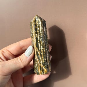 Puede incluir: Una piedra preciosa pulida y columnar con una parte superior puntiaguda. La piedra presenta un patr&oacute;n de manchas y vetas marrones oscuras, blancas y doradas. El cristal se sostiene en una mano sobre un fondo neutro.
