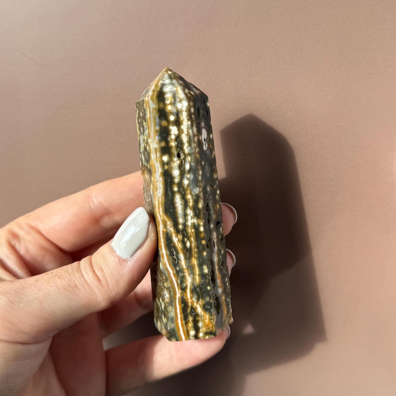 Puede incluir: Una piedra preciosa pulida y columnar con una parte superior puntiaguda. La piedra presenta un patr&oacute;n de manchas y vetas marrones oscuras, blancas y doradas. El cristal se sostiene en una mano sobre un fondo neutro.
