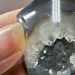 May include: A close-up of a polished, gray and clear crystal geode. The geode has a hollow center filled with small, sparkling crystals. The outer surface is smooth and reflective, with a small chip on the edge.