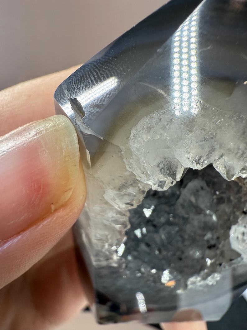 May include: A close-up of a polished, gray and clear crystal geode. The geode has a hollow center filled with small, sparkling crystals. The outer surface is smooth and reflective, with a small chip on the edge.