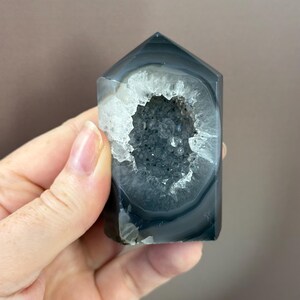 May include: A polished, rectangular agate crystal with a hollowed-out center. The crystal features bands of gray and white, with a geode-like cavity filled with sparkling, dark gray crystals. The top is angled, giving it a pointed shape.