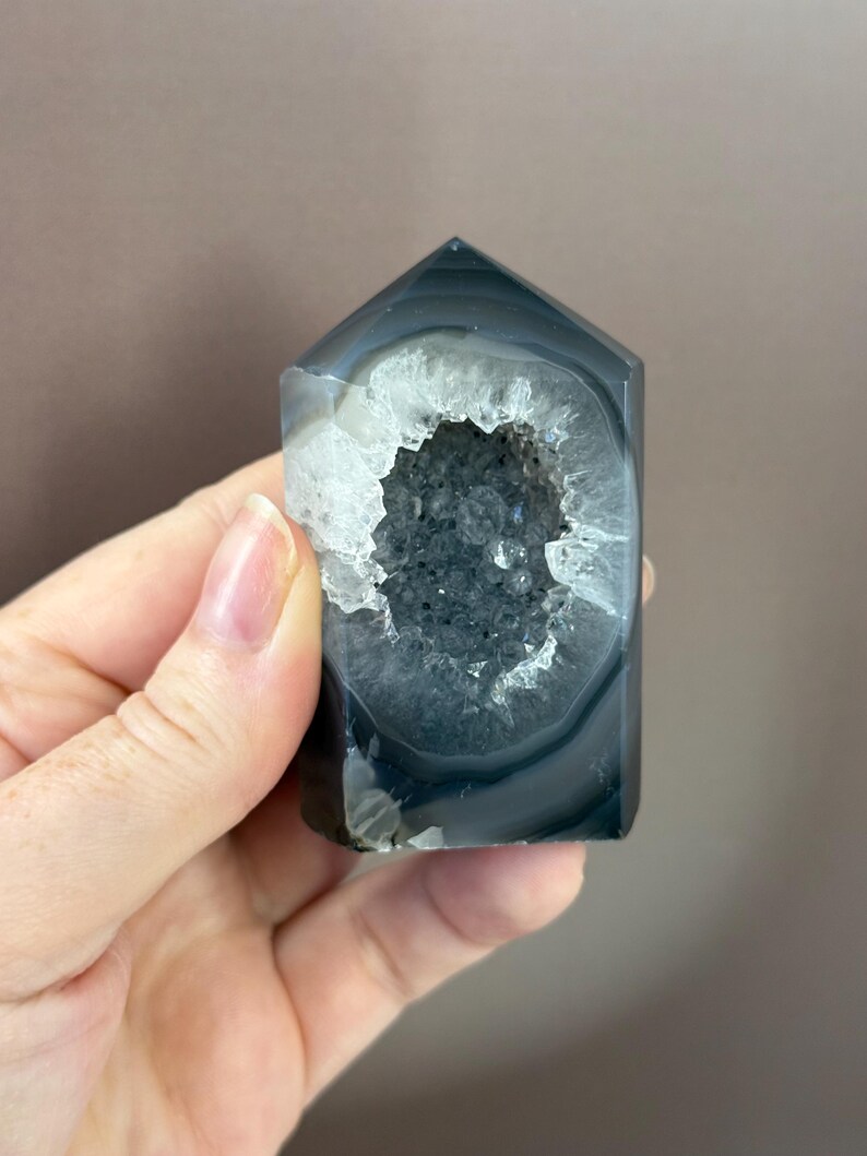 May include: A polished, rectangular agate crystal with a hollowed-out center. The crystal features bands of gray and white, with a geode-like cavity filled with sparkling, dark gray crystals. The top is angled, giving it a pointed shape.