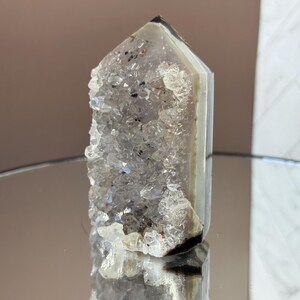 May include: A clear quartz crystal point with a rough, crystalline surface on one side and smooth, banded sides. The crystal has a natural, polished appearance and reflects light, showcasing its internal structure. The crystal is a light gray color with some dark inclusions.
