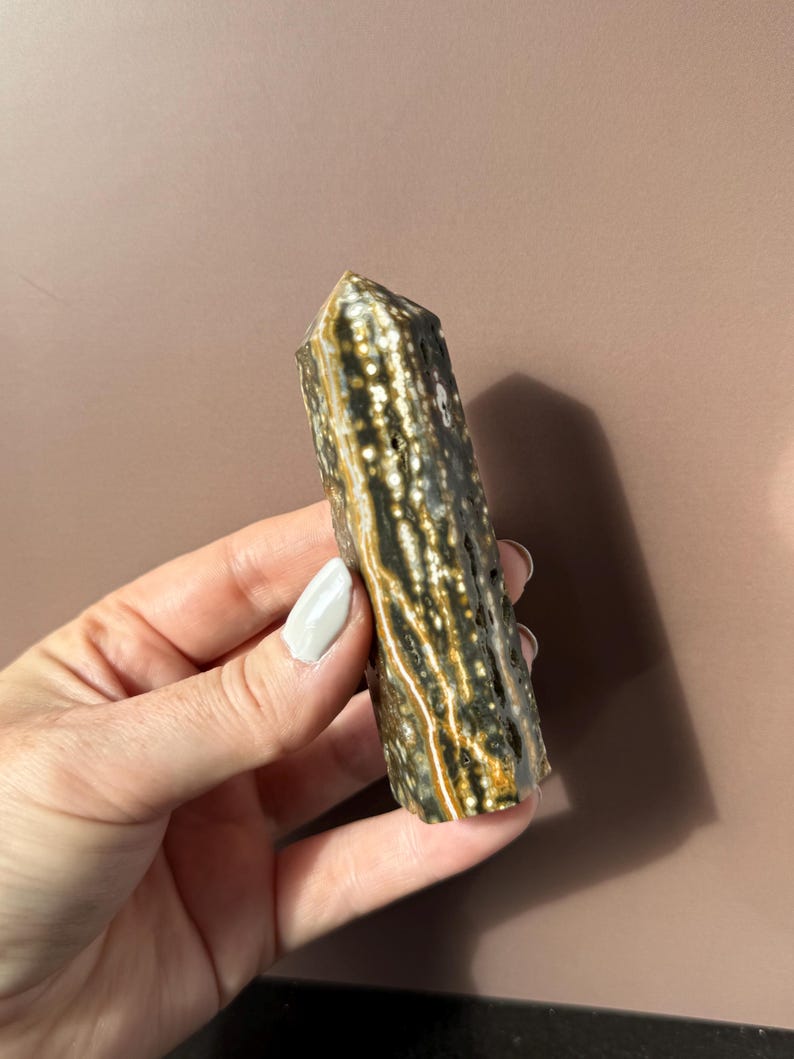 Puede incluir: Una piedra preciosa pulida y columnar con un patr&oacute;n moteado de colores verde oscuro, marr&oacute;n y crema. El cristal se sostiene en una mano sobre un fondo neutro. La parte superior tiene forma puntiaguda y facetada.