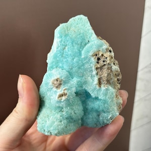 Puede incluir: Un vibrante esp&eacute;cimen mineral de color azul claro con una superficie texturizada. La piedra presenta formaciones cristalinas y &aacute;reas de inclusiones marrones y negras. La forma general es irregular, mostrando la belleza geol&oacute;gica natural.