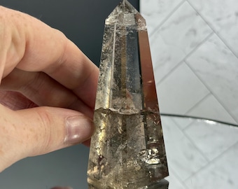 May include: A clear, faceted crystal obelisk with a pointed top. The crystal has a light brown hue and is held by a hand. The background is a mix of a dark surface and a white tiled wall.