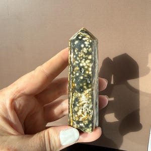 Puede incluir: Un obelisco de piedra preciosa pulida. La piedra presenta un patr&oacute;n moteado de blanco, dorado y verde oscuro. El obelisco es rectangular con una parte superior puntiaguda. La piedra preciosa se sostiene en una mano.
