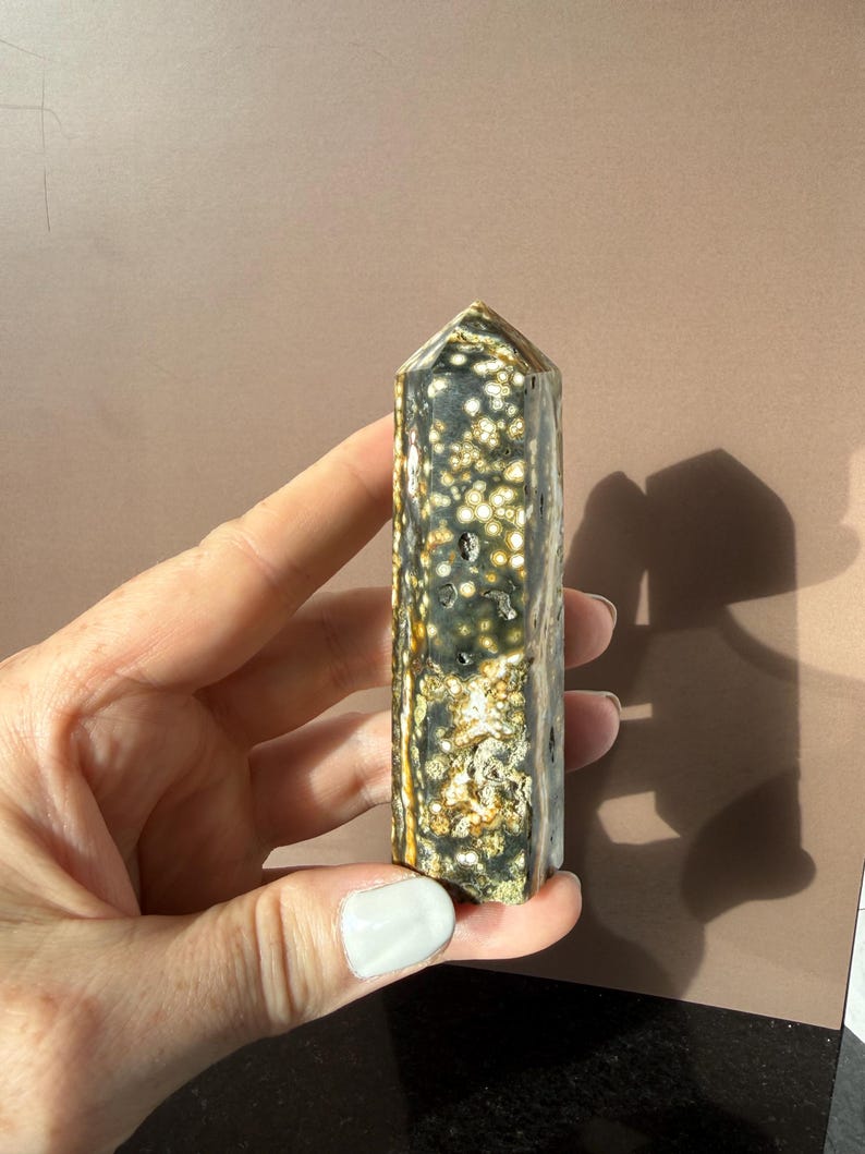 Puede incluir: Un obelisco de piedra preciosa pulida. La piedra presenta un patr&oacute;n moteado de blanco, dorado y verde oscuro. El obelisco es rectangular con una parte superior puntiaguda. La piedra preciosa se sostiene en una mano.