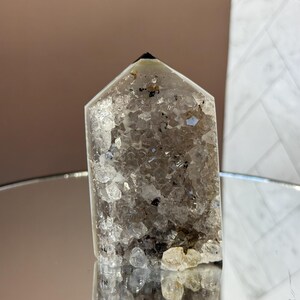 May include: A clear quartz crystal point with a faceted top. The crystal is filled with small, sparkling inclusions and has a dark tip. The crystal is displayed on a reflective surface, showcasing its natural beauty.
