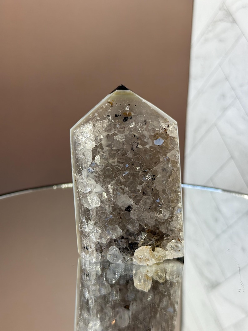 May include: A clear quartz crystal point with a faceted top. The crystal is filled with small, sparkling inclusions and has a dark tip. The crystal is displayed on a reflective surface, showcasing its natural beauty.