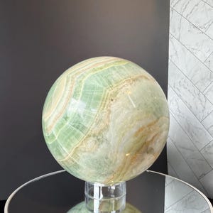 May include: A large, polished spherical stone sculpture featuring swirling bands of pale green, cream, and golden-brown mineral deposits. The smooth, rounded object rests on a small, clear cylindrical stand, showcasing natural translucent layers.