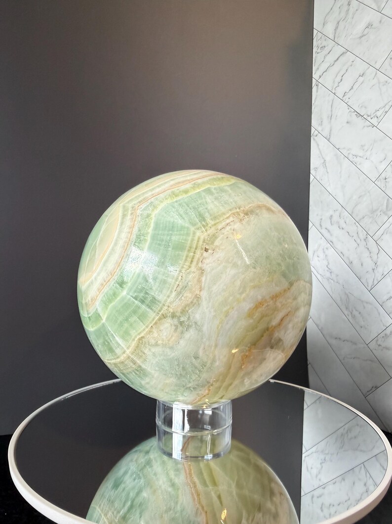 May include: A large, polished spherical stone sculpture featuring swirling bands of pale green, cream, and golden-brown mineral deposits. The smooth, rounded object rests on a small, clear cylindrical stand, showcasing natural translucent layers.