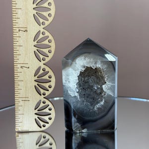 May include: A polished, gray and white agate crystal tower with a hollow, geode-like center. The crystal is approximately 4 inches tall, with a wooden ruler in the background.