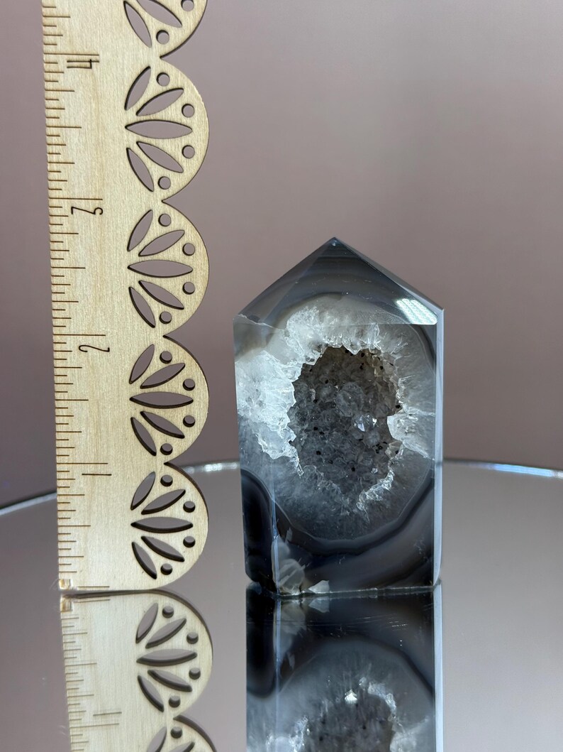 May include: A polished, gray and white agate crystal tower with a hollow, geode-like center. The crystal is approximately 4 inches tall, with a wooden ruler in the background.