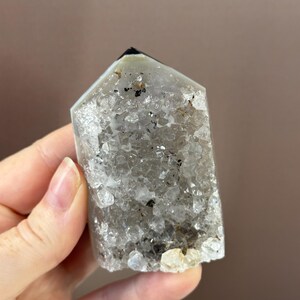 May include: A clear quartz crystal point with a faceted top and a rough, textured surface. The crystal has inclusions of black and brown minerals. The crystal is held in a hand.