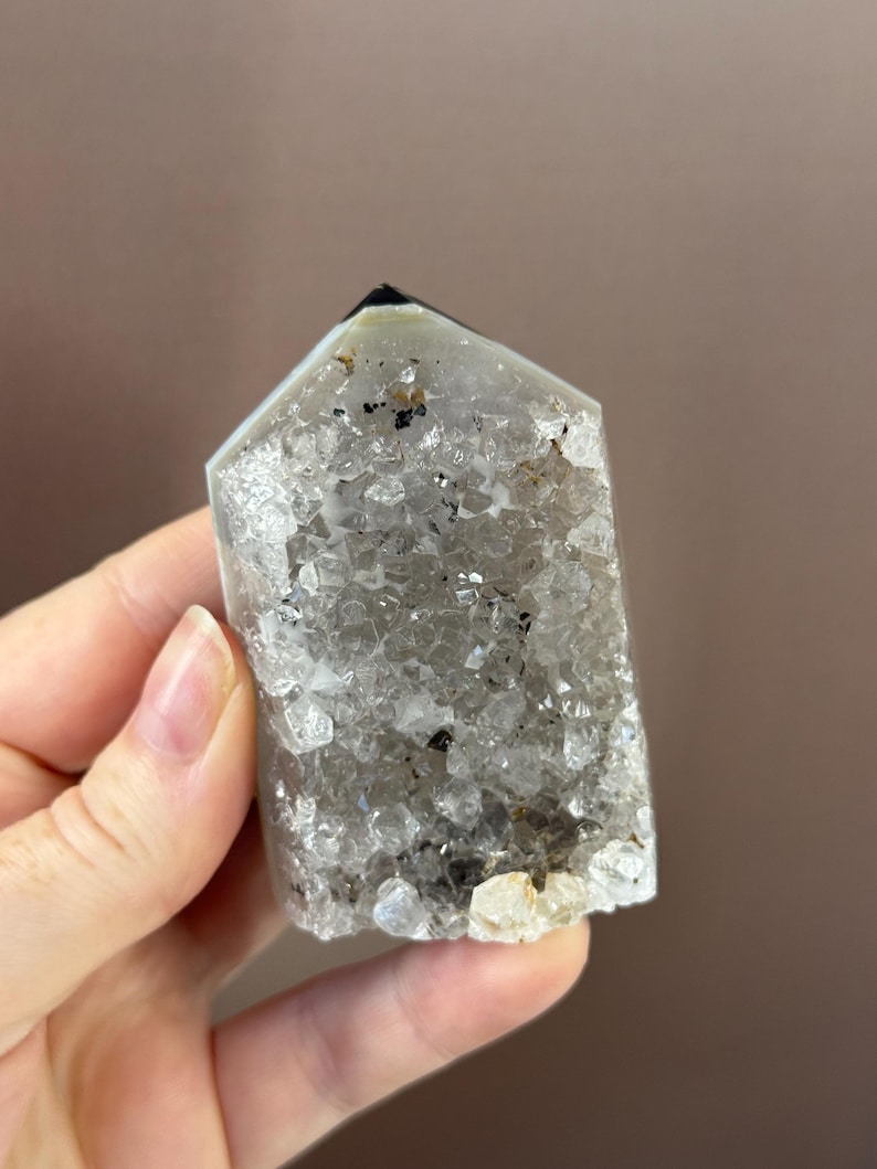 May include: A clear quartz crystal point with a faceted top and a rough, textured surface. The crystal has inclusions of black and brown minerals. The crystal is held in a hand.