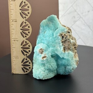Puede incluir: Un vibrante esp&eacute;cimen mineral de aragonita azul claro con una superficie rugosa y texturizada. La piedra mide aproximadamente 7.6 cm de altura, con una regla de madera para la escala. La aragonita tiene inclusiones marrones y beige.