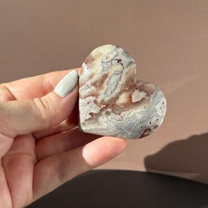 May include: A heart-shaped stone with a swirling pattern of beige, gray, and reddish-brown hues. The polished surface of the stone is smooth, and the heart is held in a hand.