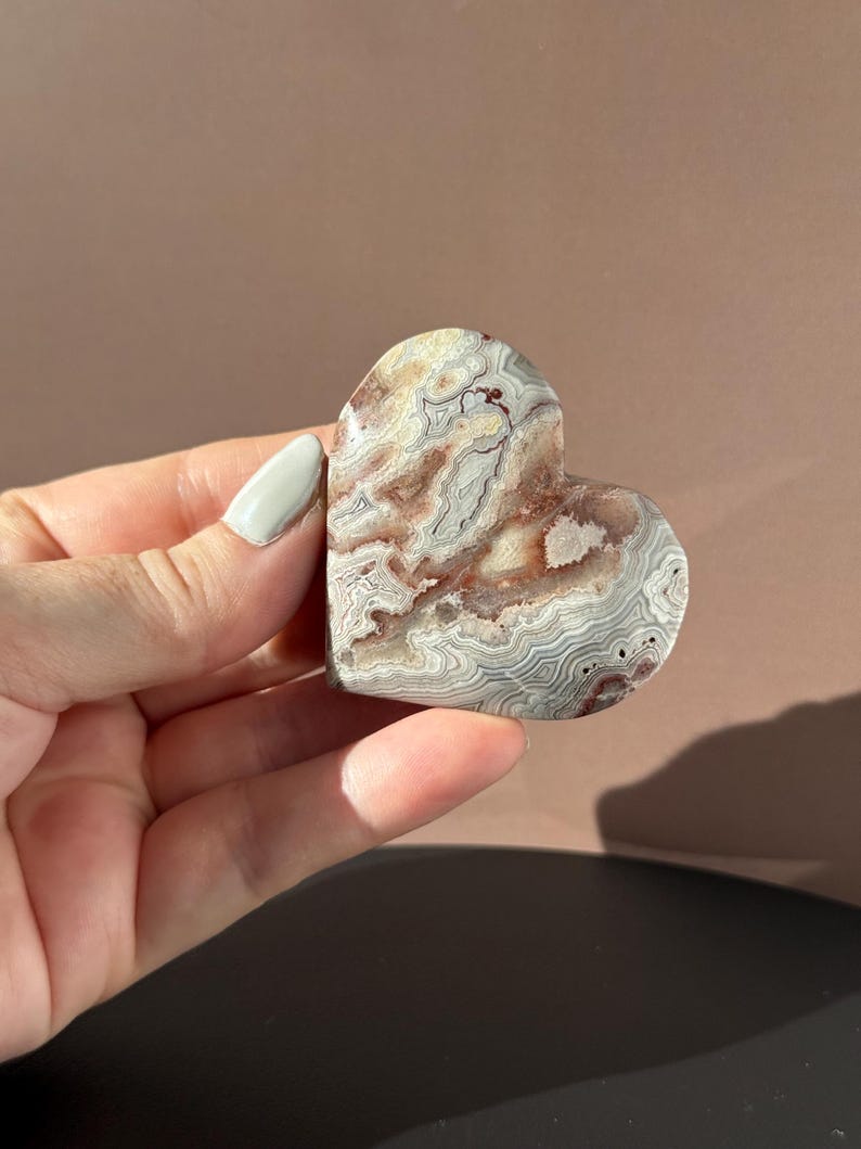 May include: A heart-shaped stone with a swirling pattern of beige, gray, and reddish-brown hues. The polished surface of the stone is smooth, and the heart is held in a hand.
