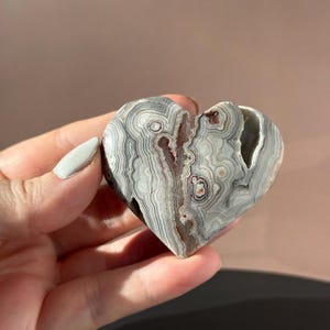 May include: A heart-shaped stone with a unique pattern of gray, white, and brown swirls. The stone has a natural, organic texture and a split down the center. The heart is held in a hand.