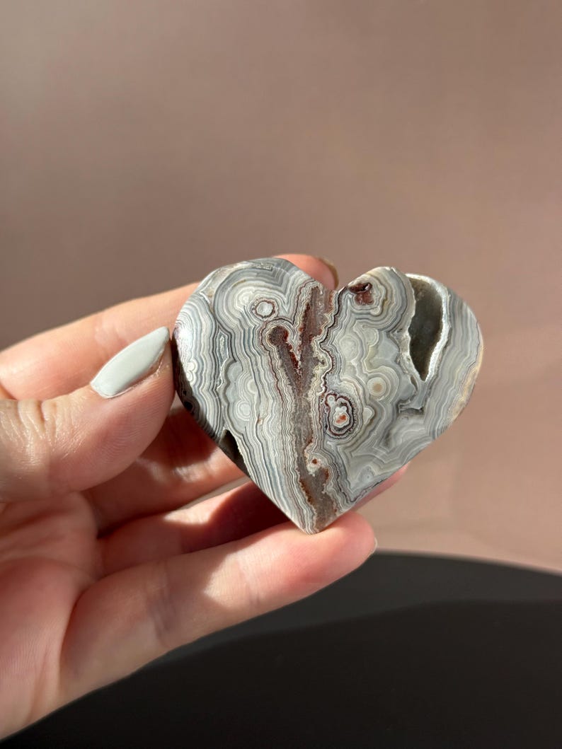 May include: A heart-shaped stone with a unique pattern of gray, white, and brown swirls. The stone has a natural, organic texture and a split down the center. The heart is held in a hand.