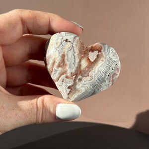 May include: A heart-shaped stone with a unique pattern of white, gray, and reddish-brown swirls. The polished surface of the stone is held in a hand, showcasing its natural beauty and intricate design. A decorative item.
