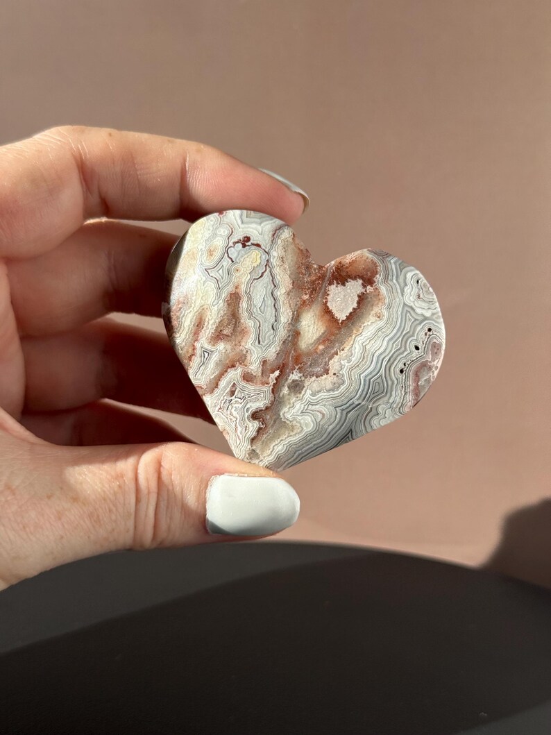 May include: A heart-shaped stone with a unique pattern of white, gray, and reddish-brown swirls. The polished surface of the stone is held in a hand, showcasing its natural beauty and intricate design. A decorative item.