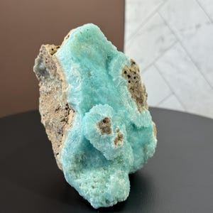 Puede incluir: Un llamativo esp&eacute;cimen mineral con una vibrante formaci&oacute;n cristalina azul claro. El mineral tiene una superficie texturizada con &aacute;reas de roca marr&oacute;n y beige. El esp&eacute;cimen se exhibe sobre un fondo neutro.