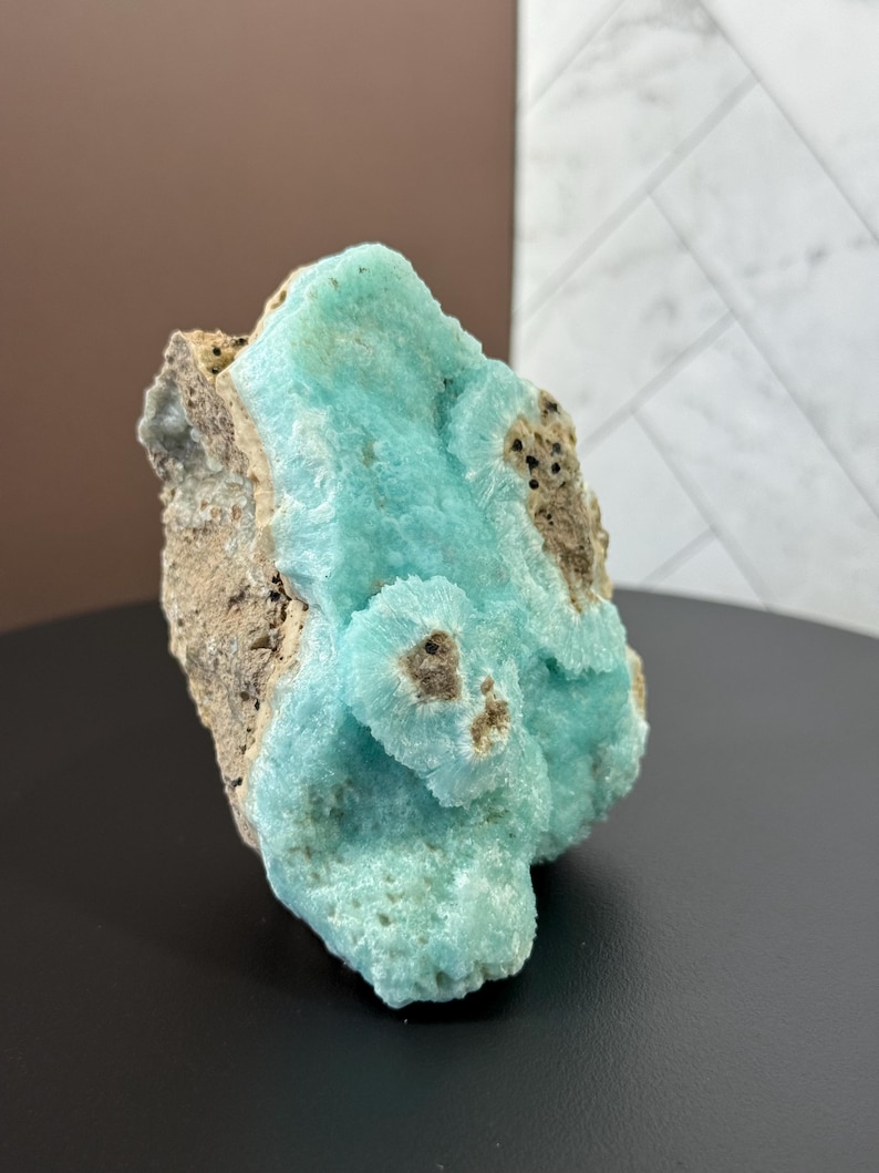 Puede incluir: Un llamativo esp&eacute;cimen mineral con una vibrante formaci&oacute;n cristalina azul claro. El mineral tiene una superficie texturizada con &aacute;reas de roca marr&oacute;n y beige. El esp&eacute;cimen se exhibe sobre un fondo neutro.