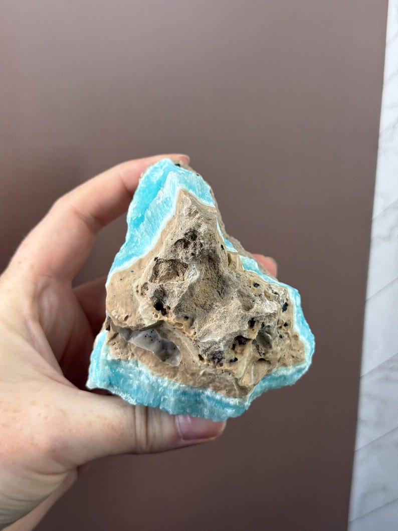 May include: A hand holds a piece of raw aragonite. The stone features a striking contrast of colors, with a vibrant turquoise blue edge surrounding a rough, tan-colored core. The aragonite has a porous texture with dark spots.