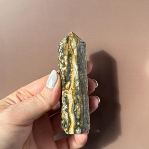 Puede incluir: Una piedra preciosa pulida, de forma columnar, con una parte superior puntiaguda. La piedra presenta una mezcla de patrones gris oscuro, negro y amarillo dorado. El cristal se sostiene sobre un fondo neutro, mostrando sus colores y texturas naturales.