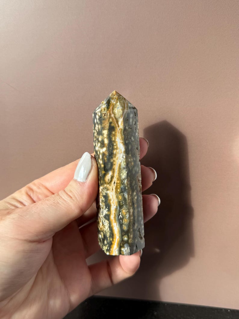 Puede incluir: Una piedra preciosa pulida, de forma columnar, con una parte superior puntiaguda. La piedra presenta una mezcla de patrones gris oscuro, negro y amarillo dorado. El cristal se sostiene sobre un fondo neutro, mostrando sus colores y texturas naturales.