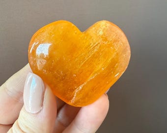 May include: A polished, heart-shaped orange gemstone. The stone has a smooth, glossy surface and a warm, inviting color. The heart is held in a hand, showcasing its size and shape. The gemstone's color ranges from a light yellow to a deep orange.