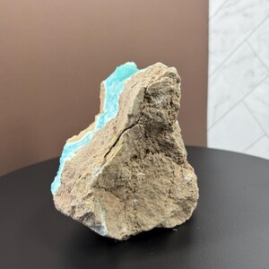 May include: A rough, natural stone specimen with a striking contrast of colors. The exterior is a muted brown, while a vibrant turquoise hue is visible within the crevices, creating a unique geological display. The stone is displayed on a black surface.