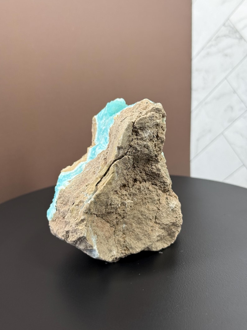 May include: A rough, natural stone specimen with a striking contrast of colors. The exterior is a muted brown, while a vibrant turquoise hue is visible within the crevices, creating a unique geological display. The stone is displayed on a black surface.