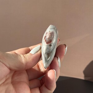 May include: A polished, teardrop-shaped stone with a layered pattern of gray and white bands. The stone features a central area of reddish-brown coloration. The stone is held in a hand.