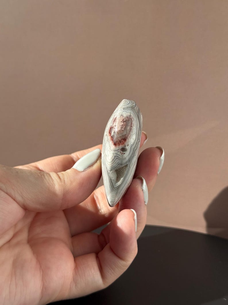 May include: A polished, teardrop-shaped stone with a layered pattern of gray and white bands. The stone features a central area of reddish-brown coloration. The stone is held in a hand.