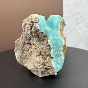 May include: A rough, natural stone specimen featuring a striking contrast of textures and colors. The stone's exterior is a mix of tan and gray, while a vibrant, crystalline formation of light blue mineral is visible.