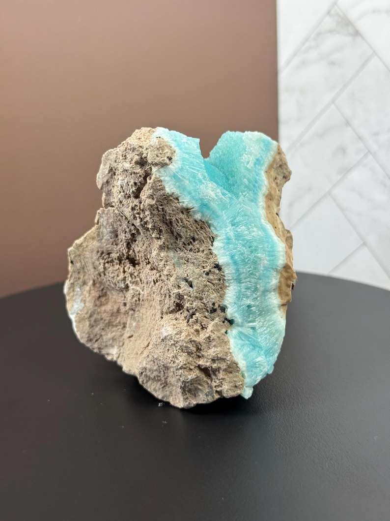 May include: A rough, natural stone specimen featuring a striking contrast of textures and colors. The stone's exterior is a mix of tan and gray, while a vibrant, crystalline formation of light blue mineral is visible.