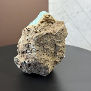 Puede incluir: Una formaci&oacute;n rocosa rugosa y texturizada con un exterior marr&oacute;n claro y un dep&oacute;sito mineral de color turquesa vibrante en la parte superior. La roca tiene peque&ntilde;as inclusiones oscuras y se asienta sobre una superficie negra. El fondo presenta una pared marr&oacute;n y una pared blanca estampada.