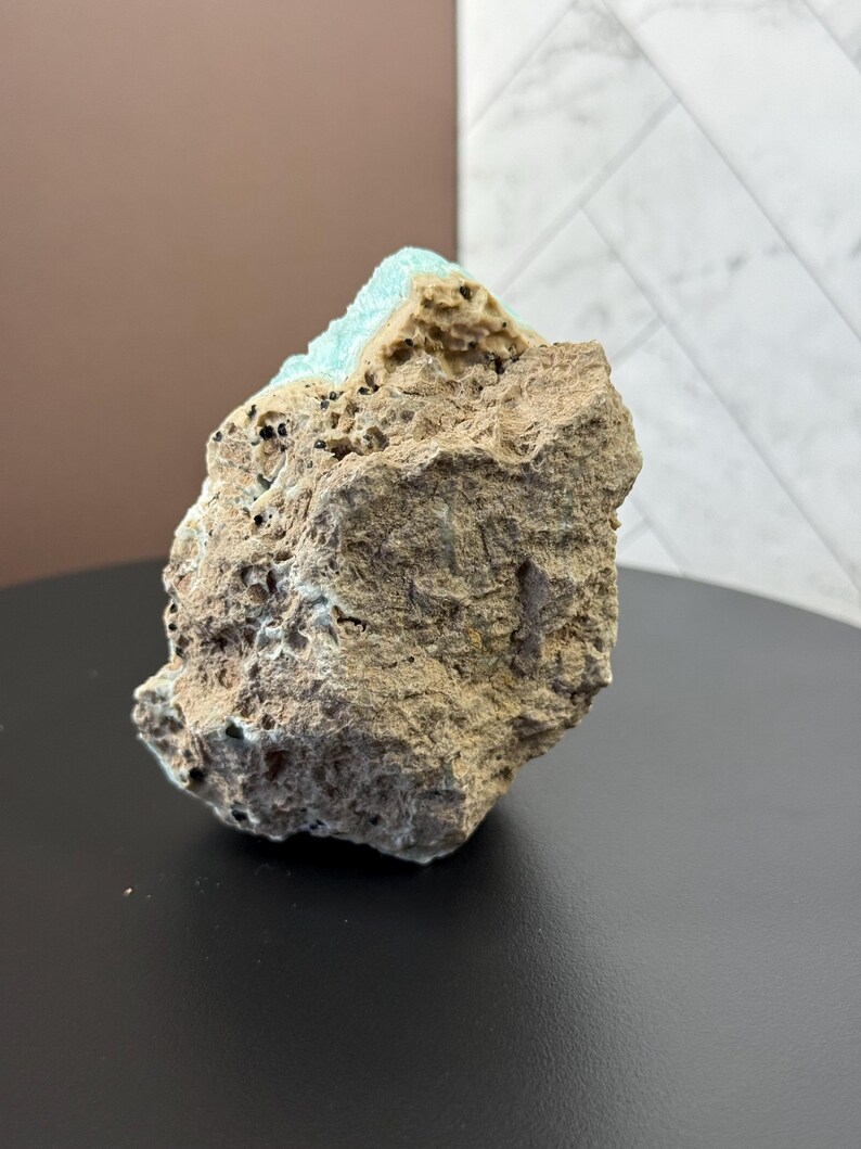 Puede incluir: Una formaci&oacute;n rocosa rugosa y texturizada con un exterior marr&oacute;n claro y un dep&oacute;sito mineral de color turquesa vibrante en la parte superior. La roca tiene peque&ntilde;as inclusiones oscuras y se asienta sobre una superficie negra. El fondo presenta una pared marr&oacute;n y una pared blanca estampada.
